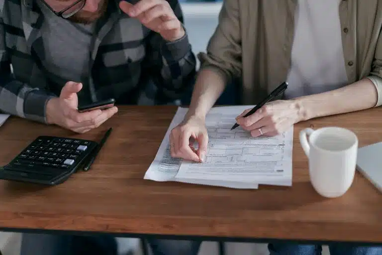 A couple sitting at a wooden table reviewing financial documents, with a calculator and a smartphone in hand, and a coffee mug nearby.